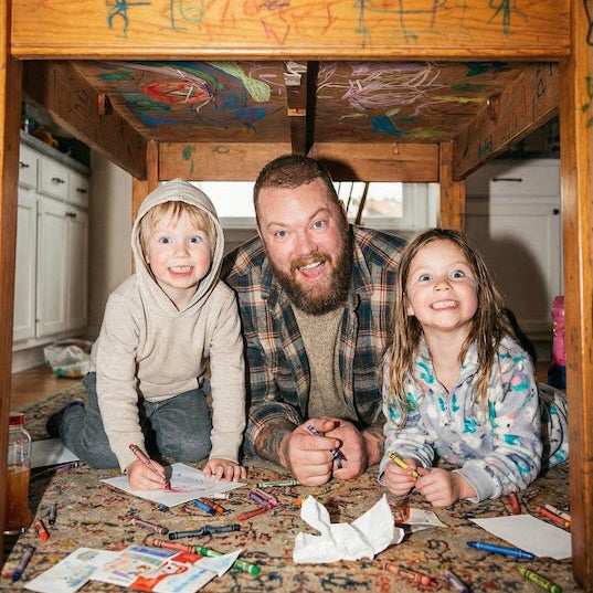 Man with two children sitting under a wooden loft bed with colorful drawings on the wall.
